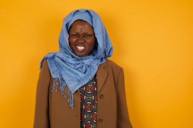 Çılgın Afrikalı Amerikalı Müslüman kadın dişlerini öfkeyle sıkar, gelen gürültüden rahatsız olur, şık kıyafetler giyer, sarı arka planda izole olur. Olumsuz duygu kavramı.