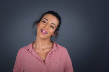 Güzel, gülümseyen esmer kadın, geniş gülümsemeli, beyaz dişleri olan, günlük pembe gömlek giyen, gri stüdyo duvarının üzerinde duran, izin gününün keyfini çıkaran.