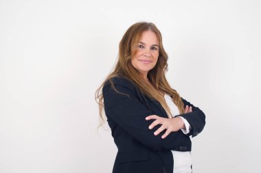 Orta yaşlı beyaz bir kadın gri duvara yaslanmış, kolları çaprazlanmış, gündelik giysiler giymiş. Kameraya bakıp gülümsüyor. Kendine güvenen başarılı bir kadın..