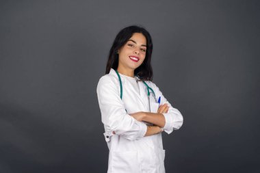 Kollarını kavuşturup tıbbi üniforma giymiş, gri duvara yaslanmış neşeli, beyaz bir doktor kadın resmi. Kameraya bakıp gülümsüyor. Kendine güvenen başarılı bir kadın..