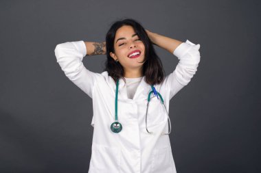 Güven ve cinsel ilişki konsepti. Çekici genç bir doktorun portresi. Kendine güvenen bir ifadeyle gülümsüyor ve ellerini başının üstünde tutuyor. Gri arkaplanda duruyor.