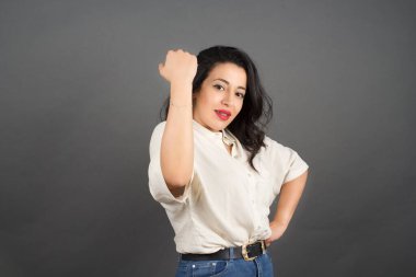 Güzel esmer kadın gelişigüzel giyinmiş yumruğunu gösteriyor. Gri arkaplanda duruyor. 