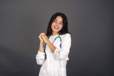 Neşeli genç doktor dişiler dişlerini sıkar, parmaklarını çapraz yapar, arzulanan dileklerde bulunur, günlük kıyafetler giyer, iyi haberleri bekler, gri duvarın üzerinde durur.