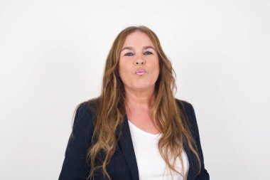 Çekici, açık renk saçlı, orta yaşlı bir kadın dudaklarını yuvarlıyor, günlük kıyafetler giyiyor, doğrudan kameraya bakıyor, memnuniyetini ifade ediyor, gri arka planda izole edilmiş. İnsanlar, vücut dili kavramı.