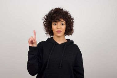 Gri duvara yaslanmış genç bir kadın bir numarayı gösterip işaret ederken kendinden emin ve mutlu bir şekilde gülümsüyor..