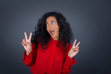 Kıvırcık saçlı, kırmızı tişörtlü, neşeli esmer kadının tek pozu iki elini de gri arkaplanda izole etmiş bir şekilde barış ya da zafer işareti yapar..