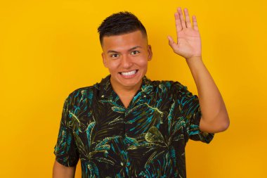 Neşeli, sempatik görünümlü, günlük kıyafetler giyen genç İspanyol bir adamın portresi. Bir elini kaldır ve merhaba de, merhaba de ve tasasız bir yüz ifadesiyle gülümse. Veda et ya da hoş geldin jesti yap..