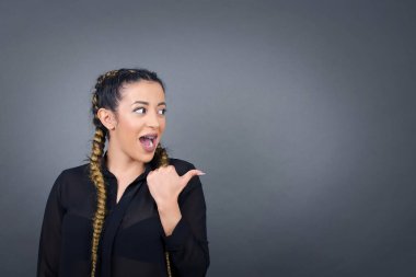 Şaşırmış esmer kadın, şaşırmış bir ifadeyle, gözlerini ve ağzını iyice açıyor, baş parmağıyla işaret ediyor, gri arka planda garip bir şey gösteriyor..