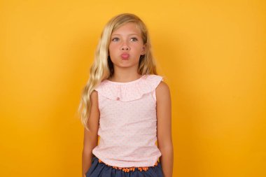 Sarışın, tatlı küçük kız sarı stüdyo arka planında dudaklarını üflüyor.  