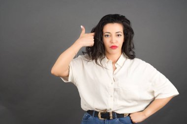 Güzel esmer kadın, gündelik giyinmiş, başparmağı havada. Gri arkaplanda duruyor. 