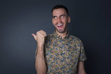 Omg konsepti. Şaşırmış beyaz bir erkek, şaşırmış bir ifadeyle, gözlerini ve ağzını iyice açıyor, baş parmağıyla işaret ediyor, gri arka planda garip bir şey gösteriyor. Reklam kavramı.