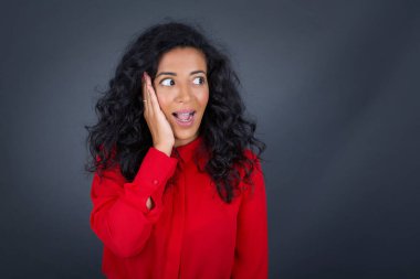 Şaşırmış, mutlu, kıvırcık saçlı, kırmızı tişörtlü, ağzı açık esmer kadın gri arka planda heyecandan yana..