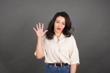 Güzel esmer kadın, gelişigüzel giyinmiş, beş parmağını gösteriyor. Gri arkaplanda duruyor. 