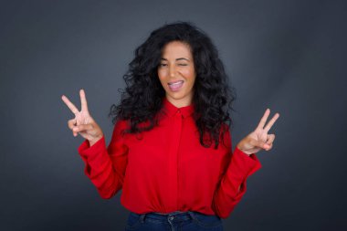 Kıvırcık saçlı, gri arka planda izole edilmiş, iyimser bir gülümsemeyle izole edilmiş, iki eliyle barış ya da zafer işareti yapan, dost canlısı görünen esmer bir kadının kapalı portresi..