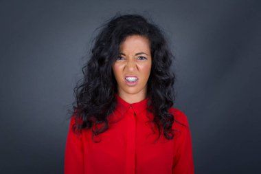 Kıvırcık saçlı, kırmızı tişörtlü çılgın esmer kadın dişlerini öfkeyle sıkar. Gri arka planda izole edilmiş gürültüden rahatsız olur. Olumsuz duygu kavramı.