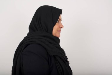 Sağlıklı tenli, siyah tesettürlü, gülümseyen Müslüman kadın profili, derin bir ifadeye sahip, açık havada yürümeye hazır, beyaz stüdyo duvarının üzerinden kopya alanı ile izole edilmiş.