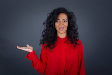 Kıvırcık saçlı, kırmızı tişörtlü, mutlu esmer kadın ifadesini hayrete düşürüyor, boş alanda bir şey gösteriyor, açık elle izole edilmiş gri arka planı var. Reklam kavramı.