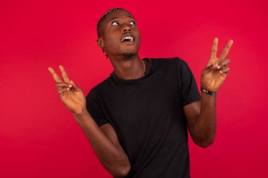 Kırmızı arka plan üzerine siyah t-shirt giyen Afro-Amerikan bir adam iki eliyle barış ya da zafer işareti yapar, rahat kıyafetler giyer, serin hisseder dişli bir gülümsemesi vardır. İnsanlar ve vücut dili.