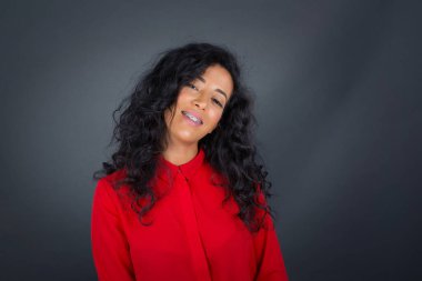 Güzel, gülümseyen esmer, kıvırcık saçlı, kırmızı gömlekli kadın gri stüdyo duvarının üzerinde duruyor..