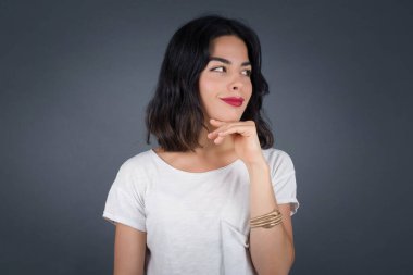 Hoş bir yüzü olan, günlük kıyafetler giyen, yan bakan, elini çenesinin altında tutan, hoş bir şeyler düşünen, gri arka planda poz veren hayalperest bir kadın..