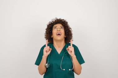 Olumlu ifadeli çekici doktor kadın, iki işaret parmağıyla işaret ediyor, tıp üniforması giyiyor.