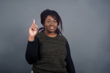  Gri duvara yaslanmış sıradan giyinmiş Afro-Amerikan kadını bir numaralı parmağını gösterip gülümserken kendinden emin ve mutlu..