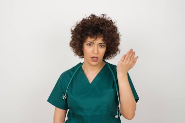 What the hell are you talking about, nonsense. Studio shot of frustrated beautiful arab doctor woman  wearing medical uniform gesturing with raised palm, frowning, being displeased and confused with dumb question over gray wall.