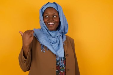 Omg konsepti. Şaşırmış Afro-Amerikan Müslüman kadın, şaşırmış bir ifadeyle gözlerini ve ağzını iyice açıyor, baş parmağıyla işaret ediyor, sarı arka planda garip bir şey gösteriyor. Reklam kavramı.