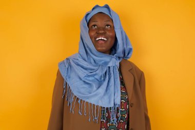 İnsanlar ve ani tepki kavramı. Şaşırmış Afrikalı Amerikalı Müslüman kadın, omuz silkiyor, yana bakıyor, arkadaşlarıyla yeni planları için mutlu ve heyecanlı. Sarı duvarda izole edilmiş..
