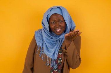 What the hell are you talking about, nonsense. Studio shot of frustrated African American muslim woman gesturing with raised palm, frowning, being displeased and confused with dumb question over yellow wall.