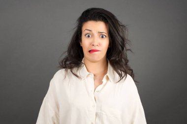 Gayri resmi giyinmiş güzel esmer kadının kafası karışmış. Gri arkaplanda duruyor.  