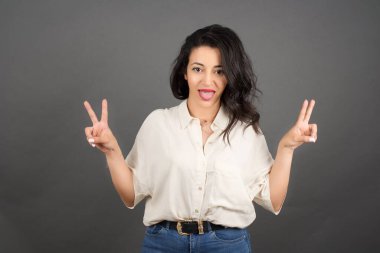 Güzelim esmer kadın zafer işaretleriyle gelişigüzel giyinmiş. Gri arkaplanda duruyor. 