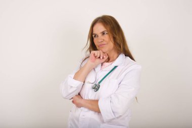 Çenesinin altında eli olan, şüpheli ve şüpheci bir ifadeyle yan bakan, şık, genç, beyaz bir kadın doktor portresi. Gri arka planda kapalı duruyor..