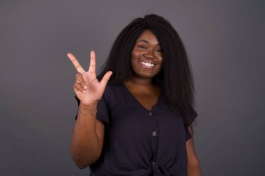 Genç Afrikalı Amerikalı kadın gri duvarın önünde duruyor. Üç numaralı parmaklarıyla işaret ediyor. Kendinden emin ve mutlu bir şekilde gülümsüyor..