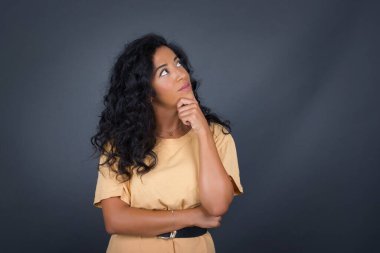 Düşünceli gülümseyen kadın portresi elini çenesinin altında tutuyor, yan bakıyor, ilgi çekici bir şey düşünüyor ya da merak ediyor, sıradan giyiniyor, gri stüdyoya karşı poz veriyor..