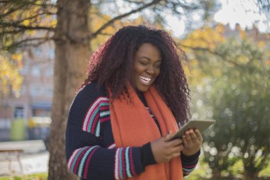 Güz parkında elinde tabletle renkli kazak giyen uzun saçlı güzel Afrikalı Amerikalı kadın.  