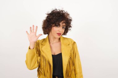 Gri duvara yaslanmış genç bir kadın, mutlu ve kendinden emin gülümserken beş numarayı gösterip işaret ediyor..