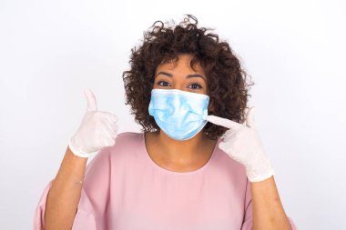 Yüz maskesi takan kadın ve beyaz arka planda baş parmağı yukarı izole edilmiş günlük kıyafetler.