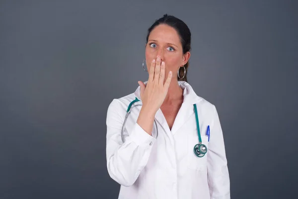 Oh! I think I said it! Close up portrait of doctor lady in white, covers open mouth by hand palm, looks at camera with big eyes isolated on bright mint background.