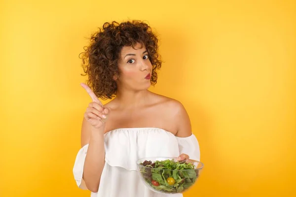 Woman holding a salad dressed casual gesturing a no sign. Closeup portrait unhappy, serious girl raising finger up saying: oh no you did not do that. Standing over yellow background. Negative emotions facial expressions, feelings.