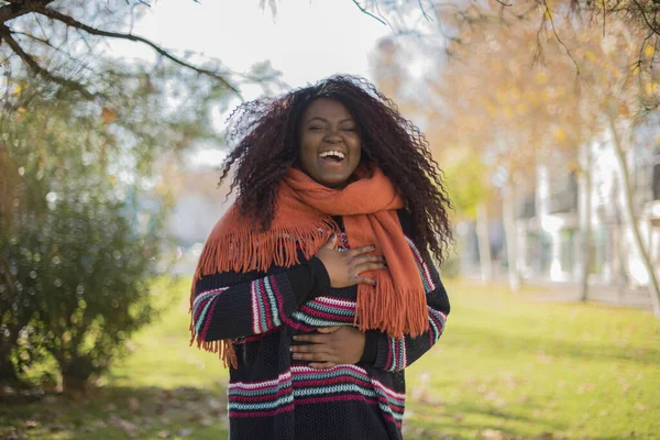 Uzun kıvırcık saçlı, renkli süveter giyen güzel Afro-Amerikan kadın parkta gülüyor.  