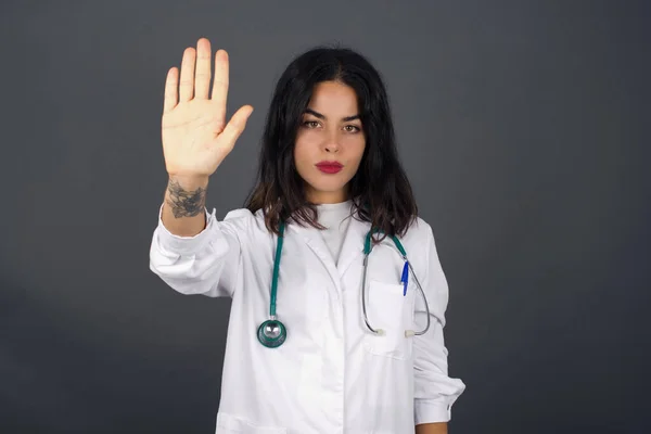 Young beautiful caucasian doctor woman doing stop gesture with palm of the hand. Warning expression with negative and serious gesture on the face isolated over gray background.