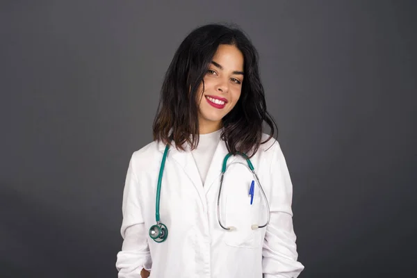 Beautiful doctor woman smiling looking to the camera wearing medical uniform and standing against gray wall.