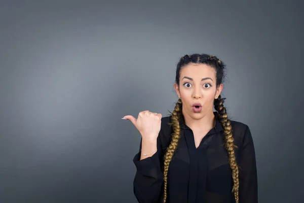 Horizontal shot of pretty young joyful shocked female points with thumb away, indicates something on blank wall isolated over gray background.