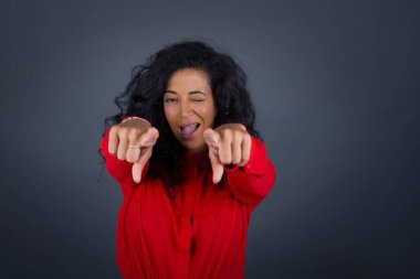 Güzel, kıvırcık saçlı, kırmızı tişörtlü, iki parmağıyla kamerayı gösteren genç, esmer bir kadın: 