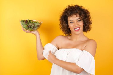 Salata salatalı güzel bir Arap kadının belden yukarı pozu kaslarını göstermek için elini kaldırıyor, zaferden emin, güçlü ve bağımsız görünüyor, kameraya olumlu gülümsüyor, arka planda duruyor. Spor konsepti.