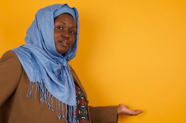 Güzel Afrikalı Amerikalı Müslüman bir kadının portresi. Sarı arka planda hoş geldin jesti yapıyor..