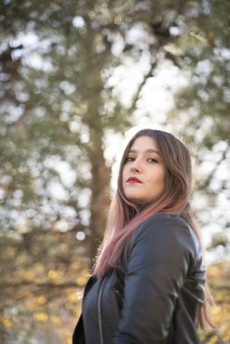 Deri ceketli beyaz bir kadın parktaki kameraya bakıyor.  