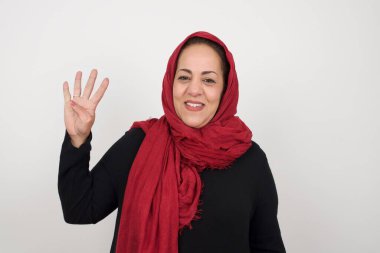 Olgun bir kadın gri duvarın önünde durmuş ve parmaklarını dört numaraya doğrultup gülümserken kendinden emin ve mutlu görünüyor. Stüdyoda poz veren başörtüsü takan orta yaşlı bir Arap kadın.