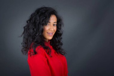 Beyaz tenli, esmer, kıvırcık saçlı, kırmızı tişörtlü, gri stüdyo arka planında duran, utangaç bir kadın.. 